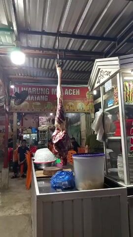 The video shows the inside of a restaurant. A large piece of meat hangs from the ceiling by a hook. Next to the meat hangs a lit glass display case selling noodles, which is in front of a stainless steel countertop. Customers are sitting at orange plastic chairs in the background. A sign reads “DI WARUNG MIE ACEH MATANG”. The restaurant has a corrugated metal roof.