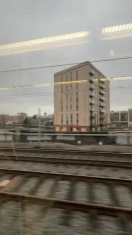 The video shows a view from a moving train. The camera is focused on the landscape passing by outside the window. The landscape consists of railway tracks, buildings of various sizes, and an overcast sky. There are no people visible in the frame. The view is somewhat obscured by the reflection of the train's interior lights on the window.