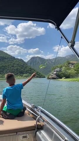 A young boy wearing a blue t-shirt and green shorts is sitting on the edge of a boat, holding a long green reed or branch. He is reaching out with it towards the water. The boat is moving through a wide, green lake, with lush, tree-covered mountains in the background. The sky is blue with scattered white clouds. The boat has a black canopy supported by metal poles. The water is calm with ripples. The perspective is from within the boat, looking out over the water.