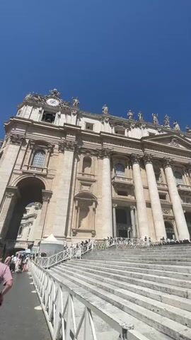 The video shows the front of St. Peter's Basilica in Vatican City, on a clear, sunny day. The camera is positioned at the bottom of the grand staircase leading up to the building, giving a low-angle view of the impressive facade. The Basilica's architecture is prominently displayed, with its large columns, statues lining the top, and a clock visible on the left side. People are standing at the base of the stairs and near the entrance, suggesting visitors are either entering or waiting to enter the Basilica. A white barrier is visible in the foreground, likely to control the flow of foot traffic. Overall, the scene captures the grandeur and scale of St. Peter's Basilica on a bright day.