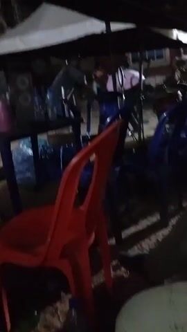 The video shows a group of people socializing outdoors at night under a white canopy. Several blue plastic chairs surround a table filled with water bottles and other items. There is one red plastic chair in the foreground. Some individuals are seated at the table, while others are standing and moving around. A speaker is visible on the table, and music is playing. The overall atmosphere appears to be casual and lively.