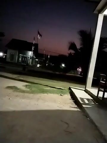 The video shows a dark outdoor scene at dusk or night. In the background, there is a small building with lights on, and two flags flying. In the foreground, there is a concrete floor or patio area with shadows cast from an unseen object to the right. The overall atmosphere is quiet and still.