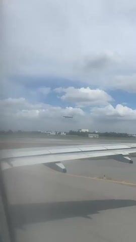 The view from an airplane window shows a runway and another airplane taking off into a cloudy sky. The wing of the plane from which the video is being shot is visible in the foreground. Buildings and trees are visible in the background between the runway and the rising airplane.