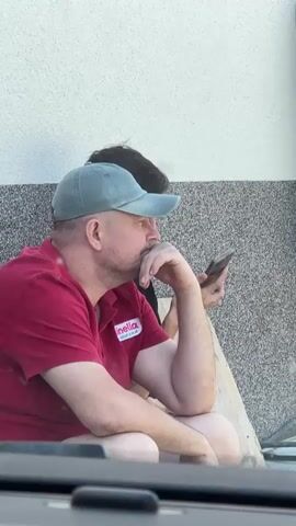 TWO MEN ARE SITTING OUTSIDE NEXT TO A WALL. THE MAN IN THE FOREGROUND IS WEARING A RED POLO SHIRT AND A LIGHT BLUE BASEBALL CAP. HE IS HOLDING A CELL PHONE IN HIS RIGHT HAND AND HIS LEFT HAND IS TOUCHING HIS CHIN. HE IS LOOKING TO HIS RIGHT. A SECOND MAN, PARTIALLY VISIBLE BEHIND HIM, IS WEARING A DARK T-SHIRT AND HAS DARK HAIR. HE IS LOOKING TO HIS LEFT. THE MEN APPEAR TO BE TAKING A BREAK OUTSIDE. THE WALL BEHIND THEM HAS A TEXTURED FINISH AND IS DIVIDED BY A HORIZONTAL LINE. THERE IS A PIECE OF WOOD OR LUMBER ON THE GROUND NEXT TO THEM.