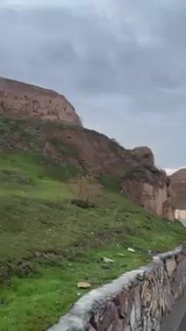 The camera slowly pans to the right, showing a road adjacent to a walking path and a dirt hill covered in green grass. The walking path is lined with a stone wall. On the far side of the road, there is a similar dirt hill topped with what appear to be ruined structures. A white vehicle drives along the road. Overhead, there are electrical lines strung between tall pylons. The sky is overcast.