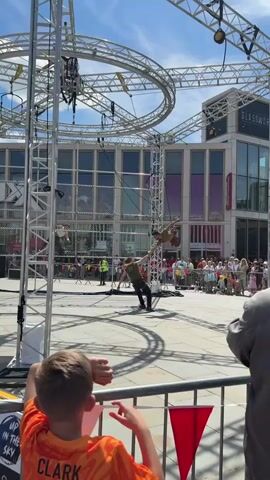 The video shows two performers suspended by ropes from a complex metal structure in a public square. One performer swings through the air while the other, standing on the ground, guides the rope. A crowd of people watches behind a metal barrier. In the foreground, a young boy in an orange t-shirt watches with his hands resting on the barrier. The sky is bright blue with light clouds. In the background, a modern building with the word "Glassworks" visible on its facade. The overall scene suggests an aerial acrobatic performance taking place outdoors.