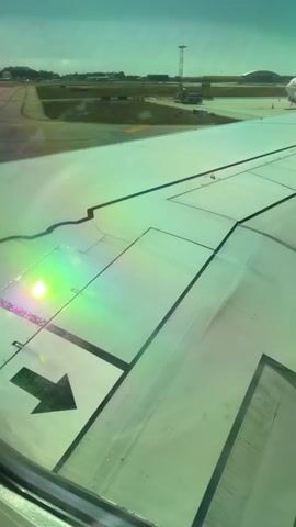 The video shows a view from an airplane window, looking out at the wing and the tarmac of an airport. The wing is white with black markings, including a large black arrow pointing forward. There are reflections and iridescence on the wing, possibly due to sunlight hitting the window. In the background, there are airport buildings, a control tower, and another airplane parked on the ground. The sky is a hazy blue-green. The camera appears to be stationary, offering a static view of the scene. There are no people visible in the video, and no actions are taking place other than the implied movement of the airplane itself. The overall impression is one of being on the ground at an airport, preparing for or having just arrived from a flight.