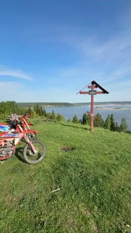 The video shows a scenic view from atop a grassy hill, with a wooden cross in the foreground and a wide body of water in the background.  To the left, a parked red dirt bike is partially visible. The sky is blue with scattered white clouds, indicating a clear day. Evergreen trees dot the landscape leading down to the water's edge. The cross, painted red, stands prominently on the hill, with a small, dark roof-like structure covering the top. The water appears calm, with islands and landmasses visible in the distance.