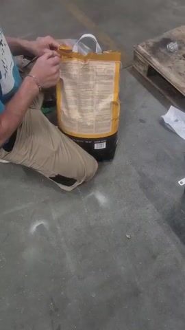 A person wearing tan pants and a teal t-shirt is kneeling on a concrete floor, opening a large yellow and black bag. The bag has handles on top. The person's hands are visible as they pull open the top of the bag. To the right of the bag, a wooden pallet is visible. The environment appears to be an industrial or warehouse setting.