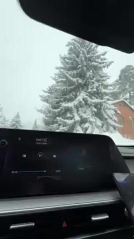 From inside a car, the view through the windshield shows a snow-covered landscape of trees and houses. The car seems to be slowly moving. A large infotainment screen shows a song playing, “Had To” by Kevin Gates. The screen also displays time elapsed and remaining. The driver’s phone is also visible.