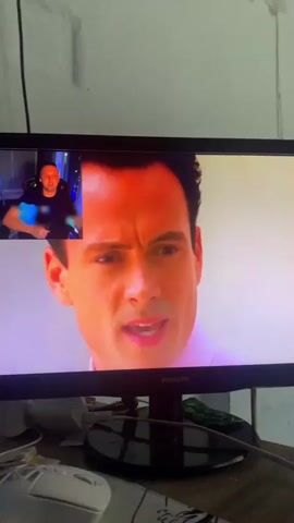 The video shows a computer monitor displaying a scene from a movie or TV show. A smaller window in the upper left corner shows a man watching the screen. The main scene on the monitor features a man in a pink shirt and then a woman in a bikini. Later, it shows a man with sunglasses and a woman laughing. The monitor is on a desk with a computer mouse and cables.
