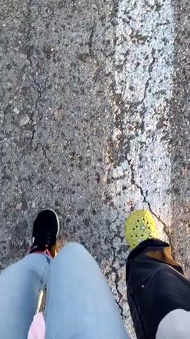 The video captures a low-angle view of two pairs of feet walking on a cracked asphalt surface. The person on the left is wearing light blue jeans and black sneakers with pink accents. The person on the right is wearing yellow Crocs and black pants. The ground appears to be a roadside, with dry grass and weeds visible on the left side. In the distance, a white car is parked on the side of a paved road that stretches towards the horizon under a clear blue sky with scattered clouds. The sun is low in the sky, casting long shadows and illuminating the scene with warm, golden light, suggesting it is either early morning or late afternoon. The overall atmosphere is relaxed and casual.