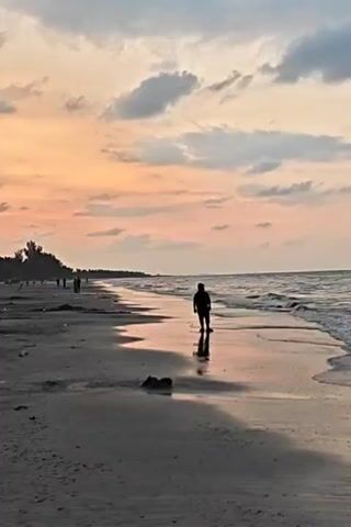 The video captures a serene beach scene at dusk, under a sky painted with hues of orange and grey. A silhouette of a person stands on the wet sand near the water's edge, their reflection visible on the smooth surface. In the distance, several other figures can be seen strolling along the beach. The beach appears to have dark sand and some debris scattered on it. The waves gently lap against the shore. The scene evokes a sense of solitude and natural beauty.