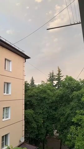 A video captures a view from a window, looking out at a building and trees under a cloudy sky. The building, a light brown multi-story structure with white-framed windows, is on the left side of the frame. The trees, lush and green, dominate the right side and the background. The sky above is a mix of gray clouds with hints of light breaking through, creating visible sunbeams. Power lines are visible crisscrossing the upper portion of the frame. The camera pans slightly, shifting the perspective of the building and trees.