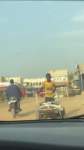The video shows a view from inside a car on a dirt road in a town. A young man is pulling a cart filled with what appears to be food or produce towards the car. He is wearing a yellow t-shirt and black shorts. Other vehicles, including motorcycles and a three-wheeled auto rickshaw, are also on the road. Buildings and shops line the sides of the road under a clear sky.