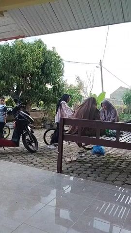 A group of people are standing in the tiled-floor patio area of a house. A young boy is on a small motorbike, and a woman in a hijab and light-colored jacket is on a bicycle. She is pushing the bike, which has a cover on the back. Two women are sitting on a bench next to the bike. There are two motorbikes parked nearby, and a tree and field can be seen in the background. The sky is overcast.
