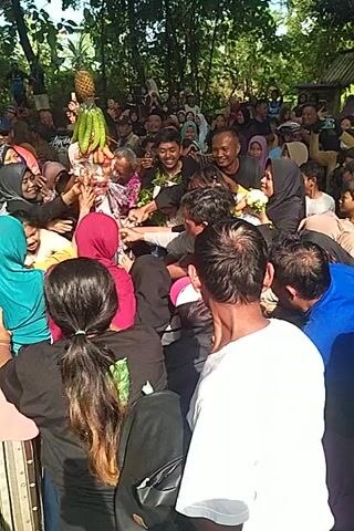 Here is a description of the video:

**Overall:** The video shows a dense crowd of people gathered around a central point where a variety of fruits and vegetables are placed. 

**People:**
*   There is a large crowd of people, mostly adults.
*   Many women are wearing hijabs (headscarves) in various colors.
*   Some individuals are reaching towards the central display of fruits and vegetables, possibly trying to grab them.
*   The expressions on people's faces suggest excitement or eagerness.

**Objects:**
*   The central display includes pineapples, bananas, cucumbers, and other fruits/vegetables arranged on a platter or tray.
*   Some individuals are wearing casual clothing such as T-shirts and jackets.
*   A few are wearing hats.

**Actions and Interactions:**
*   People are crowded closely together, reaching towards the central display.
*   There is a sense of activity and movement as individuals try to get closer to the fruits and vegetables.
*   The overall atmosphere suggests a communal event or a distribution of goods.

**Environment:**
*   The setting appears to be outdoors.
*   Trees are visible in the background.
*   The lighting suggests it is daytime.