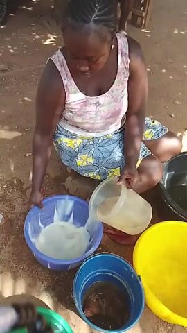 A woman sits cross-legged on the ground, working with several buckets and containers. She is wearing a floral tank top and a patterned blue and yellow skirt. Her hair is braided. In front of her are five buckets in various colors: blue, yellow, black, green, and a blue one with a darker substance inside. She is pouring liquid from a clear container into a blue bucket that contains a whitish liquid. The ground is a mix of dirt and sand, and there is a wooden stool with someone sitting on it in the background. The overall setting appears to be outdoors, possibly in a yard or a village.