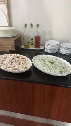 This video shows a buffet-style food spread, likely in a restaurant or event setting in Çanakkale Merkez, Turkey. The scene is well-lit, suggesting daytime. Several white platters are arranged on a dark countertop, filled with various salads and dishes.

In the foreground, three large oval platters are visible. The one on the left contains a creamy salad with diced vegetables, the middle platter holds a similar creamy salad with chunks of red pepper and other ingredients, and the platter on the right is filled with a green salad, possibly with herbs. Further back, more platters of food are lined up, including what appears to be a potato salad, a mixed vegetable salad, and other dishes in white serving bowls.

Behind the main buffet line, a wooden tiered stand holds more food items. A large white bowl sits on the top tier. Three glass bottles, one containing yellow liquid (likely oil), another with a yellow citrus-based liquid, and a third with a red liquid (possibly a dressing or sauce), are placed on a white tray. A stack of white plates is visible to the right.

In the background, a wall features a framed piece of art with an abstract design. Further down the buffet, a row of green apples is displayed in a white platter, followed by watermelon slices and cucumber slices. Additional dishes with red ingredients, possibly tomato-based salads, are also present.

The overall impression is that of a diverse and abundant buffet offering a variety of culinary options. The setting appears to be indoors, with a clean and organized presentation of food. No specific emotions are conveyed, but the abundance of food suggests a pleasant dining experience. No visible text is present in the video.