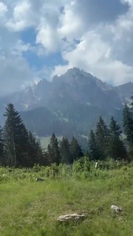 The video shows a mountainous landscape with lush green grass and trees in the foreground. In the background, there are towering, rugged mountains with some trees and greenery on their slopes. The sky is partly cloudy, with patches of blue and white clouds. The camera pans slightly, providing a panoramic view of the scenic environment. There are no people or distinct actions depicted.