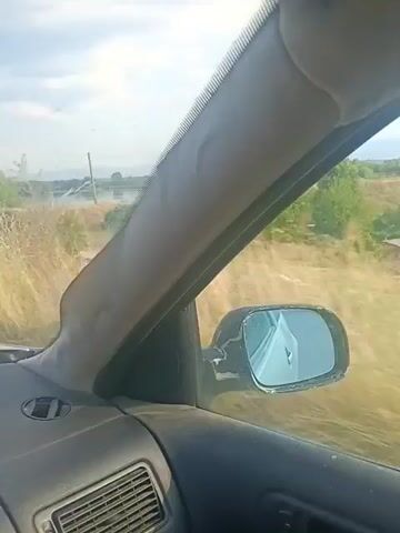 The video shows a view from inside a car, looking out the passenger window. A side mirror is visible in the foreground, reflecting the sky and the interior of the car. The landscape outside is dry and dusty, with tall, brown grass and some scattered trees. In the distance, there is a body of water, possibly a lake or pond, and a small building with a red roof. A white car is parked near the building. The sky is cloudy.