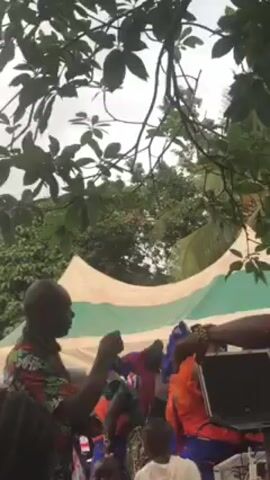 Here's a description of the video:

The video shows a lively outdoor event with music and dancing. A group of women in matching blue and orange outfits are dancing in front of a white tent. A DJ is seen behind a table with a laptop, managing the music. Several other people are around, some dancing, some watching, and some working. The environment appears to be a yard or outdoor venue, with trees visible overhead.