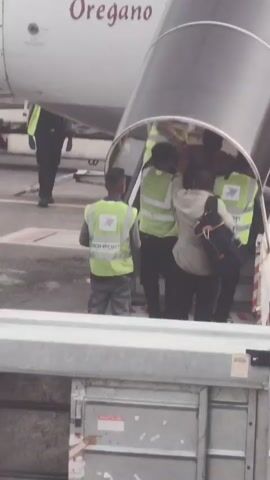 At an airport tarmac, people are boarding a plane via a mobile staircase. A person in a grey uniform with a yellow vest stands to the left, facing away from the staircase. Several individuals in yellow vests with "AIRPORT" printed on them are guiding passengers up the stairs. Some passengers are visible ascending the stairs, one carrying a duffel bag. The aircraft has "Oregano" printed on its side. In the foreground, a metal baggage cart is visible.