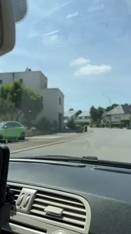 The video is shot from inside a car, looking out through the windshield. The car is driving on a paved road in a residential area. The houses are modern and appear to be well-maintained, with light-colored facades. There are trees and bushes lining the road, and streetlights are visible. The sky is blue with a few white clouds. The dashboard of the car is visible in the foreground. The car appears to be turning a corner. There are garbage bins visible on the side of the road.