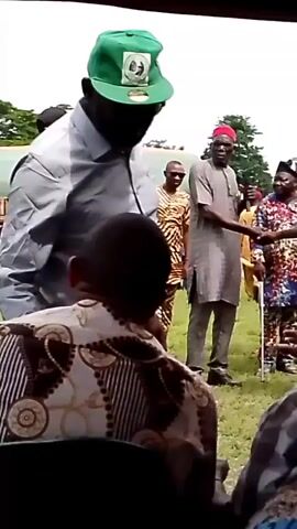 The video shows an outdoor gathering with several people. In the foreground, the back of a person wearing a patterned shirt is visible. In the middle ground, a man in a light-colored shirt and green cap is facing away from the camera, looking towards a group of people. To his right, a man in a grey traditional outfit and a red cap is shaking hands with another person, who is partially obscured. Behind them, other individuals are present, some wearing colorful patterned clothing. The environment appears to be a grassy area, possibly a park or an open field, with some trees and tents in the background. The lighting suggests it is daytime.