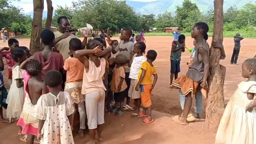 A group of about 20 children, both boys and girls, mostly of African descent, are gathered in a dirt clearing. They are clustered around a man who is holding a clear plastic bag. The man is gesturing with his other hand as he speaks to the children. Most of the children are looking at the man, listening attentively. Some children are looking around the clearing. A few trees are scattered around the clearing, and in the background, there are rolling hills. Other people are present in the background. The day appears to be sunny.