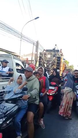 A street scene with a large sound system mounted on a truck in the background. Many people are present, some on motorcycles, others standing or walking. The atmosphere appears to be festive, possibly a parade or celebration. The time is daytime, with clear skies.