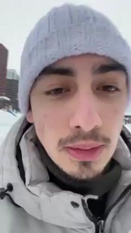 Here is a description of the video.

A young man with dark hair and a small mustache stands in a snowy area. He wears a light grey beanie and a puffy, light grey jacket. Behind him, modern high-rise buildings are visible against an overcast sky. The man says "Bye bye." The camera then tilts upward to show the skyscrapers from a lower angle.