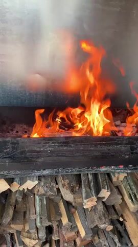 The video shows a close-up view of a fire burning in a metal smoker or grill. Flames of various sizes and intensities flicker and dance among pieces of wood. Below the fire pit, a stack of neatly cut firewood is visible, suggesting preparation for continued burning. The background is a brushed metal surface, likely part of the smoker's interior. The lighting is dominated by the warm glow of the fire.