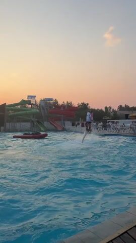 The video captures a sunset scene at a water park. A person on a jetpack is suspended in the air above a wave pool, spraying water. In the background, there are water slides, trees, and a crowd of people gathered around the edge of the pool. One person is in the water, near the edge of the pool, and another person appears to be holding onto them. A red inflatable raft is floating in the water. The sky is a gradient of orange and pink, indicating the setting sun.