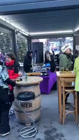 The video shows a gathering of people in an outdoor bar or patio area. A DJ with a saxophone is performing music behind a set of equipment on a purple-covered table. Several people are standing around, some holding drinks, watching the performance. There are small wooden tables with chairs and a larger table fashioned from a wooden barrel. The area is covered by a pergola-style roof with lights. The background shows more people and buildings, suggesting an outdoor event or market.