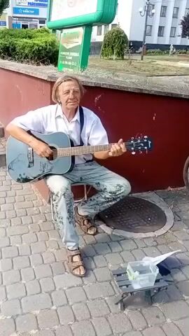 A man with blonde hair, wearing a white shirt, suspenders, and light-colored pants with a floral pattern, is sitting on a stool on a cobblestone sidewalk and playing an acoustic guitar. He is positioned in front of a dark red wall. To his right, there is a manhole cover and a small makeshift table with a clear plastic container holding some items, likely for donations, and a white paper. In the background, there is a green advertising board, some bushes, and a building. The scene appears to be outdoors in daylight.