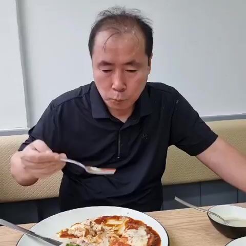 A middle-aged man with thinning hair, wearing a black polo shirt, is sitting at a table and eating from a white plate with a fork. He is in the process of taking a bite of food from the plate, which contains a dish with sauce and what appears to be meat and cheese. He is looking down at the plate as he eats. The background shows a light-colored wall and a beige seating area.