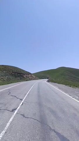 The video shows a paved road curving through a grassy, hilly landscape under a clear blue sky. The road has white lines marking its lanes. On the right side of the road, there is a concrete barrier with black and white stripes. A small vehicle is visible in the distance on the road. The hills are covered in green grass, and some bare earth is visible on the left side of the road. The camera appears to be mounted on a moving vehicle, as the perspective of the road and surroundings shifts.