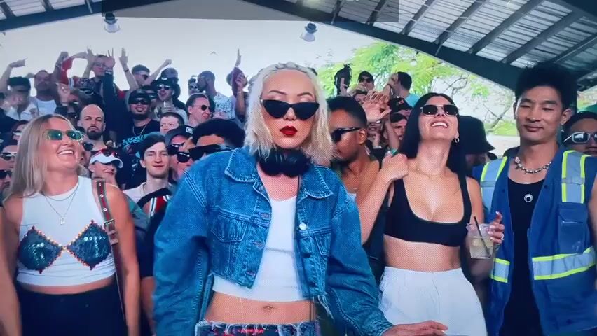 A DJ is performing at an outdoor party. She is a blonde woman with sunglasses, a white top, a cropped denim jacket, and headphones around her neck. The crowd behind her is dancing and cheering. Some are wearing sunglasses and holding drinks. The party seems lively and energetic. The location is a covered outdoor space.