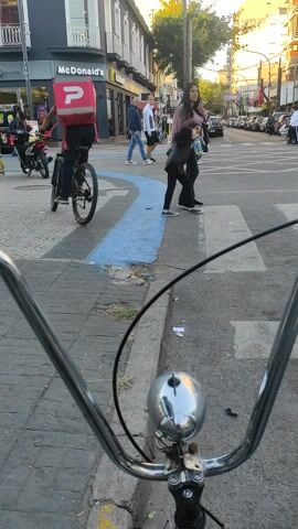 The image presents a first-person perspective from someone on a bicycle, looking down a street in Lanús Este, Argentina, during the daytime. In the immediate foreground, shiny chrome bicycle handlebars and a reflective chrome bell are prominently featured, along with a black cable.

The scene unfolds on a paved street with a sidewalk or pedestrian area to the left. Several people are visible in the midground. Two women stand near a blue painted line on the ground, one in a pink/purple top and dark pants, the other in a dark jacket and pants, both appearing to be observing the street. Further back, another individual in a blue shirt is seen. A delivery person on a bicycle, equipped with a prominent red delivery box (likely for food), is stopped or moving slowly in front of a commercial building. Other bicycles and motorbikes are parked nearby on the sidewalk.

The background is dominated by urban architecture. A McDonald's restaurant is clearly visible on the left side, identified by its distinct yellow "McDonald's" signage. Beyond it, other buildings line the street, with trees and foliage appearing above them. On the right side of the street, vehicles are parked parallel to the curb. The sky is bright, suggesting clear weather. The overall impression is a busy, typical street scene in an urban or suburban setting, with daily activities taking place.