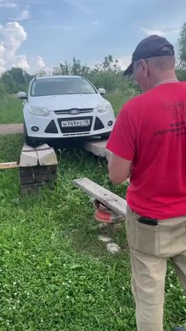 A white Ford Focus is partially elevated on wooden blocks and what appears to be a metal ramp with wheels. A man wearing a red t-shirt and tan pants stands in the foreground, facing away from the camera, looking at the car. The car is in a grassy area with trees and a cloudy sky in the background. The license plate of the car is visible, displaying H749PC 18. The man's t-shirt has a logo and text on the back, which appears to be related to sports and tourism. The lighting suggests it is daytime.