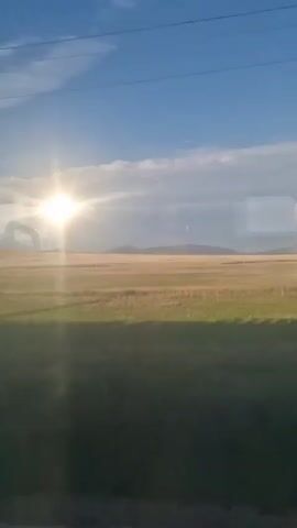 The video shows a view from a moving vehicle, likely a train, looking out at a rural landscape. The sun is bright and high in the sky, casting a glare on the window. The landscape consists of fields of grass and crops, with distant hills or mountains. There are utility poles and wires visible along the tracks. The reflection of the interior of the vehicle is also visible on the window, obscuring parts of the outside view. The video appears to be filmed in daylight, with a clear blue sky and some scattered clouds.