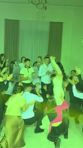 A group of people are dancing at a party. In the center of the frame is a woman in a white dress, possibly a bride. She is smiling and has her arms outstretched. She is surrounded by other guests, some of whom are also dancing. The room appears to be a ballroom or event hall, with curtains in the background. Lighting effects are present, casting colorful glows on the scene. The overall atmosphere is celebratory.