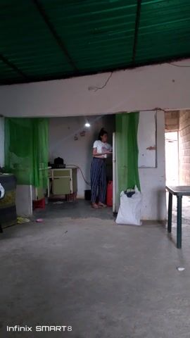 The video shows a woman indoors drinking from a pink cup. The woman is wearing a white t-shirt and striped pants. She has her hair pulled back. The room appears to be unfinished, with concrete floors and walls that are partly brick. There is a green curtain in the background. There is a table in the foreground. The video is recorded in vertical format.