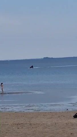 The video shows a calm beach scene. The foreground is sandy beach, and in the distance is a calm, blue body of water. There is a jet ski moving across the water, leaving a white wake. Two children, a girl and a younger child, are walking in the shallow water near the shoreline. To their left, a rock barrier stretches out into the water. The sky is overcast and light blue.