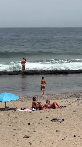 The media file captures a serene beach scene in Arona, Spain, under a hazy, likely warm, daytime sky. The foreground is dominated by a sandy beach littered with small dark stones. A vibrant blue beach umbrella stands to the left, casting a shadow on a pink towel where two young women are sunbathing. One woman is lying down, the other is sitting up, both in bikinis, enjoying the sun. Their relaxed postures suggest a peaceful day of leisure.

Beyond the beach, a natural pool or calm inlet is visible, separated from the open ocean by a dark, rocky barrier. In the water, a person walks towards the shore. Further out, in the ocean, a couple stands near the rocky edge, seemingly interacting. The waves are moderately sized, indicating a gentle surf. A lone surfer is visible further out in the ocean, riding a wave. The muted colors of the ocean and sky suggest it might be midday or slightly overcast. The overall atmosphere is tranquil and idyllic, depicting a classic beach vacation scene. There is no visible text in the media file.