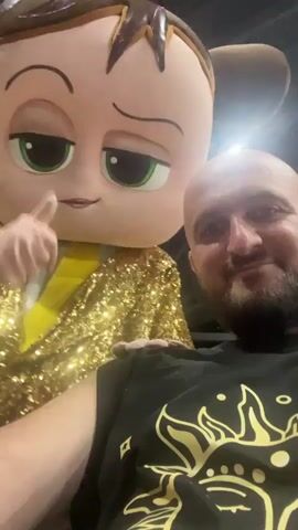 A man in a black t-shirt with a sun design is taking a selfie with a person in a Boss Baby costume. The Boss Baby character is wearing a sparkly gold jacket and has one hand on the man's shoulder, the other hand is on the man's face, pulling his cheeks to make him smile. The man is smiling, looking at the camera, and has a beard. The background appears to be indoors with dark walls and some lighting.