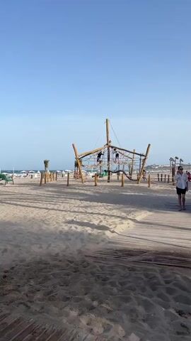 This is a vertical video of a sandy beach scene on a clear, sunny day. The time of day appears to be late afternoon or early evening, as indicated by the long shadows cast on the sand and the soft lighting. The location is Alicante, Spain.

In the foreground, a wooden boardwalk leads onto the beach, with sand covering much of the frame. To the left, a green park bench is visible, and further back, beach umbrellas and lounge chairs are set up. A prominent feature on the beach is a large, wooden rope-climbing playground structure, resembling a ship's mast and sails, with several children playing on it.

In the middle ground, a young girl in a bikini is standing near an outdoor shower, appearing to wash herself off. Other people are scattered across the beach, some walking, some sitting, and some interacting near the water. The sea is visible in the background, with gentle waves and a few people swimming or wading. Palm trees are interspersed along the beach, adding to the tropical atmosphere.

On the boardwalk in the foreground, there are white post-like structures with a symbol of a crossed-out cigarette, indicating a no-smoking zone. A woman in a white shirt and dark shorts is walking away from the camera on the boardwalk. The overall mood is relaxed and leisurely, typical of a beach vacation.