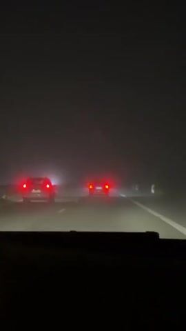 A low-angle interior shot from a car shows two red taillights of cars ahead on a foggy highway at night. The visibility is low, and the red taillights appear blurred. The road ahead is barely visible, with the fog obscuring the distant view. A faint blue light is visible in the distance on the left side of the frame. The camera is slightly shaky, indicating motion. The dashboard of the car is visible in the bottom portion of the frame, showing a yellow illuminated symbol.
