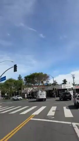 The video shows a street intersection on a sunny day with light clouds in the sky. The camera pans to the left, following the traffic flow. Several cars, including a white van and a black SUV, are visible on the road. Buildings line the street, with trees planted along the sidewalks. There are traffic signals and crosswalks visible at the intersection. A "No Left Turn" sign is also present. The scene appears to be in a suburban or urban environment, with a mix of commercial and residential buildings.